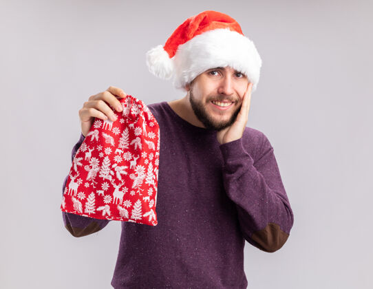 年轻人穿着紫色毛衣 戴着圣诞帽的快乐的年轻人手里拿着红包 手里拿着礼物 站在白色的背景下 看着镜头 开心地微笑着包开心礼物