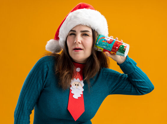 成人自信的成年白人女性 戴着圣诞帽 打着圣诞领带 拿着纸杯 靠近耳朵 橙色背景 留有复印空间圣诞老人抱着杯子