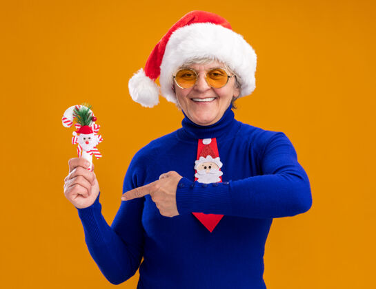 举行戴着太阳眼镜 戴着圣诞帽 系着圣诞领带的老妇人高兴地站着 指着橘黄色背景上的糖果手杖 手杖上留有复制空间圣诞老人老人分