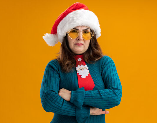 眼镜戴着太阳眼镜 戴着圣诞帽 打着圣诞领带 双臂交叉 站在橙色背景上 留有复印空间的未经许可的成年白人妇女站着圣诞老人不放