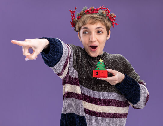 穿着令人印象深刻的年轻漂亮女孩戴着圣诞花环手持圣诞树玩具和日期看 并在紫色的墙上孤立的一面指着花环印象深刻圣诞节