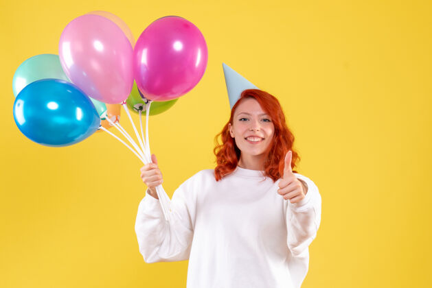可爱黄色墙壁上手持可爱彩色气球的年轻女子的正视图年轻的女性情感年轻