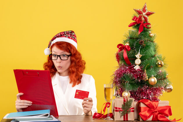桌子正面图：女医生坐在桌子后面 手里拿着黄色背景的银行卡 还有圣诞树和礼品盒卡片节日人