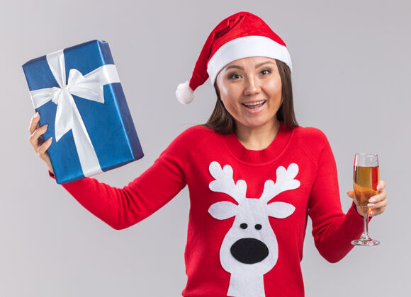 礼物笑容满面的年轻亚洲女孩 戴着圣诞帽 穿着毛衣 手里拿着礼品盒 白色背景上隔着一杯香槟香槟盒子圣诞节