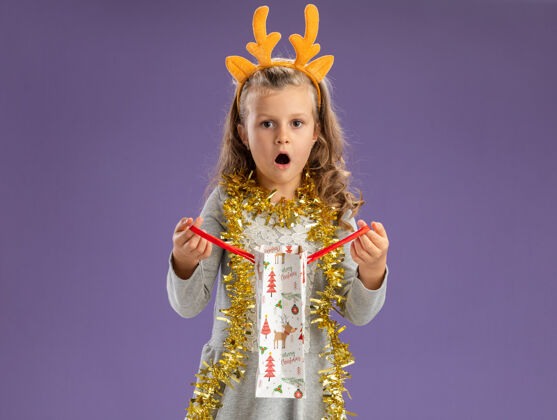 女孩惊讶的小女孩戴着圣诞发箍 脖子上戴着花环 手里拿着蓝色背景的礼品袋箍抱花环