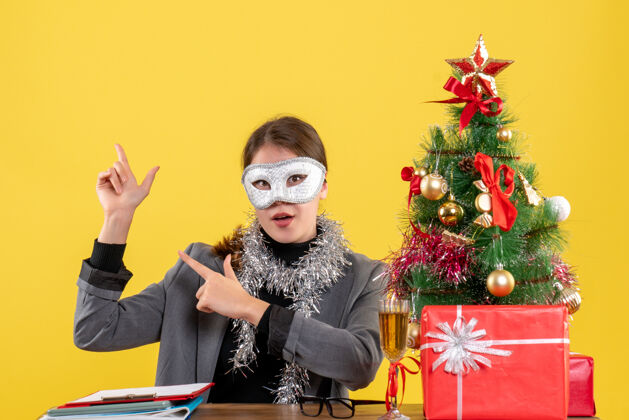 面具正面图戴着面具的年轻女孩坐在桌旁 手指着圣诞树和礼物鸡尾酒装饰礼物年轻女孩
