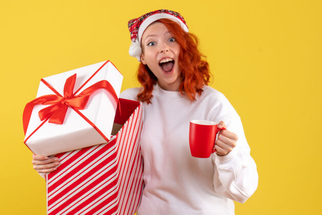 微笑黄色墙上年轻女子手持圣诞礼物和一杯茶的正面图节日礼物礼物