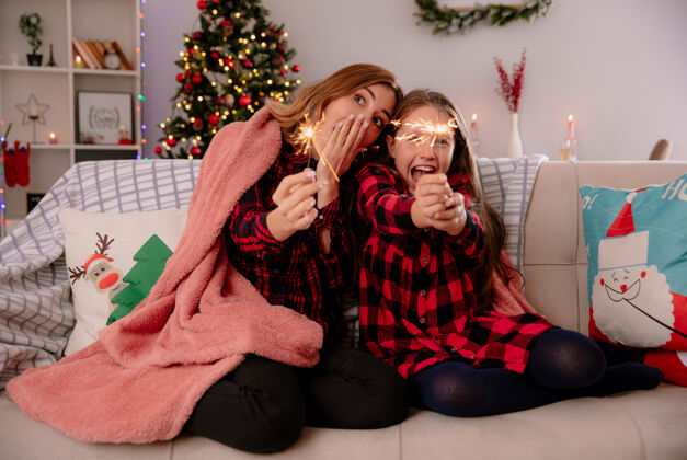 兴奋兴奋的母亲和女儿抱着闪闪发光的毯子坐在沙发上 享受圣诞节在家里的时光沙发女儿毯子