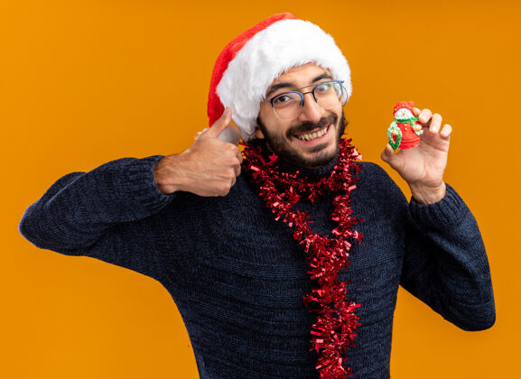 年轻微笑着的年轻帅哥戴着圣诞帽 脖子上戴着花环 手里拿着玩具 在橙色背景上孤立地竖起大拇指帽子家伙花环