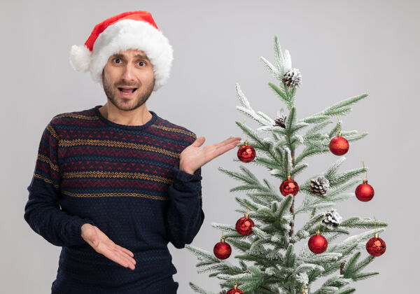 指着令人印象深刻的年轻白人男子戴着圣诞帽站在圣诞树旁 用手指着隔离在白色墙上的圣诞树站着靠近戴着