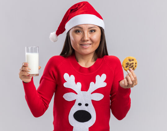 女孩微笑的亚洲女孩戴着圣诞帽 穿着毛衣 手里拿着一杯牛奶和饼干 背景是白色的微笑饼干毛衣