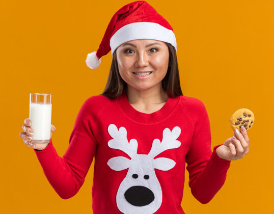 牛奶笑容满面的年轻亚洲女孩戴着圣诞帽 穿着毛衣 手里拿着一杯牛奶和饼干 隔离在橙色的墙上女人玻璃穿着