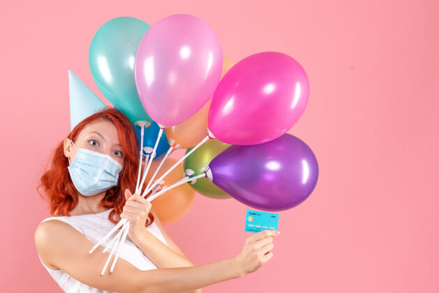 气球前视图中的年轻女子拿着五颜六色的气球在无菌面具上粉红色的墙壁Covid乐趣无菌