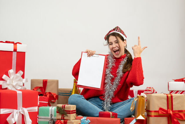 礼物快乐可爱的女孩 戴着圣诞帽 手指朝上 拿着文件坐在白色的礼物旁边文件椅子人