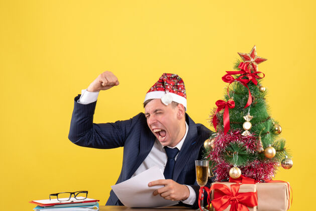 树前视图兴高采烈的男子显示获胜的姿态坐在圣诞树附近的桌子上 黄色的礼物礼物西装男人