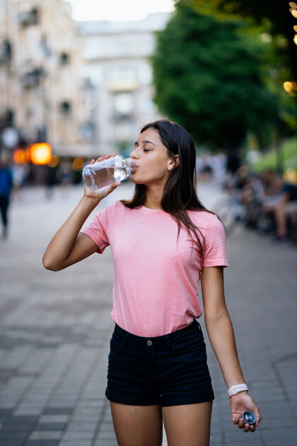 女性美丽的年轻女孩在城市街道上用一瓶水喝瓶子城市背景