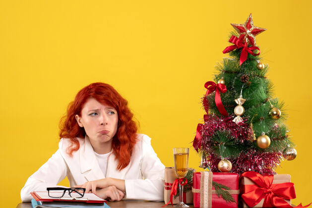 黄色女工人坐在桌子后面 手里拿着圣诞礼物 树在黄色的树上感到悲伤树花女性