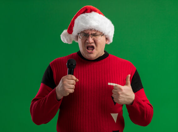 眼镜自信的成年男子戴着眼镜 戴着圣诞帽 拿着麦克风 看着相机 在绿色背景上孤立地竖起大拇指圣诞快乐圣诞拇指