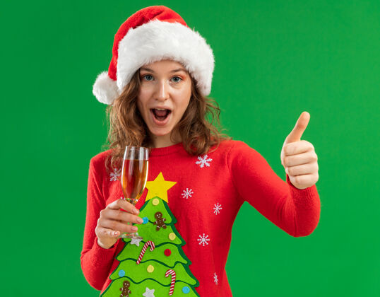 微笑身穿红色圣诞毛衣 戴圣诞帽的年轻女子手持一杯香槟看着摄像机 兴高采烈地微笑着竖起大拇指站在绿色背景上开心年轻人帽子