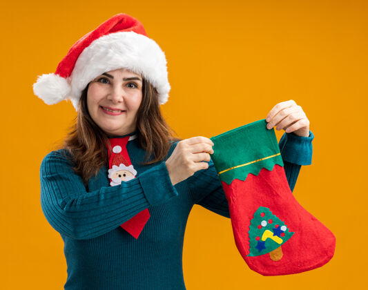 成人微笑的成年白人妇女 戴着圣诞帽 打着圣诞领带 手里拿着圣诞长袜 橙色背景 有复制空间帽子微笑抱着
