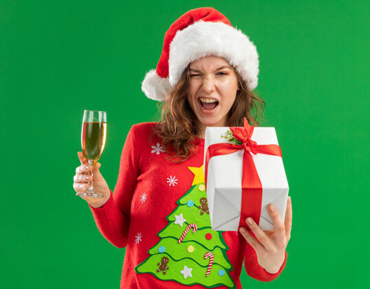 眨眼穿着红色圣诞毛衣 戴着圣诞帽 手持香槟和礼物的快乐年轻女子站在绿色背景下 看着镜头 眨着眼睛 愉快地微笑着礼物玻璃杯圣诞老人