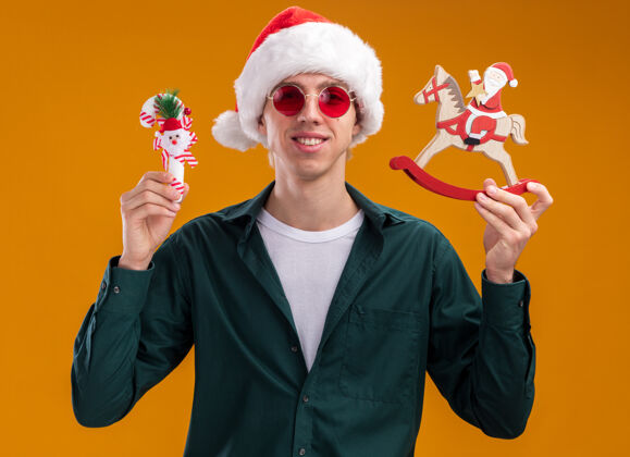 帽子带着圣诞帽戴着眼镜的年轻金发男子微笑着拿着摇摆木马上的圣诞老人雕像和甜美的藤条装饰看着橙色背景上孤立的相机圣诞老人金发马
