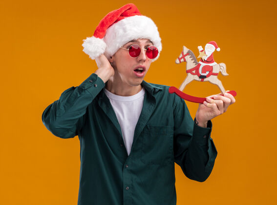 年轻令人印象深刻的年轻金发男子戴着圣诞帽和眼镜拿着圣诞老人在摇摆木马雕像看着相机保持手在头上孤立的橙色背景头手保持