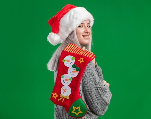 长袜年轻的金发女郎穿着冬季毛衣 戴着圣诞老人帽 肩上扛着圣诞长袜 站在绿色的背景下 快乐而快乐地微笑着圣诞老人站着毛衣