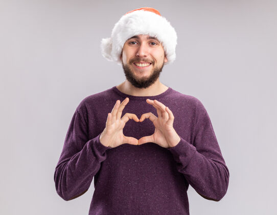 微笑穿着紫色毛衣 戴着圣诞帽的快乐的年轻人看着相机 站在白色的背景上 用手指做着开心的手势圣诞老人手势心