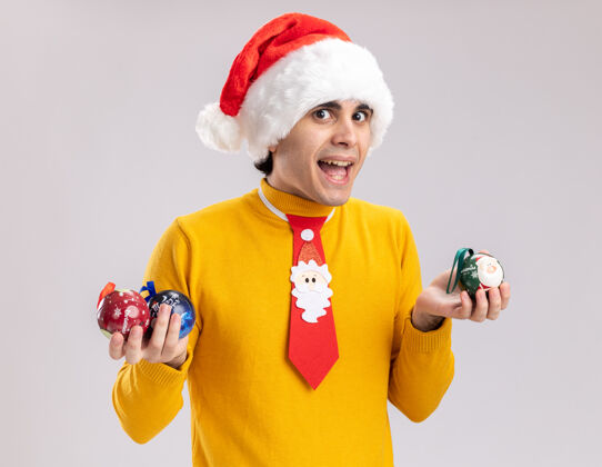 快乐身着黄色高领毛衣 戴圣诞帽 手持圣诞球 站在白色背景下 喜气洋洋地看着摄像机的快乐年轻人帽子站着举行