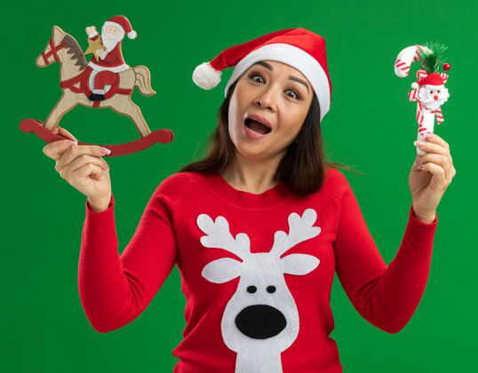 圣诞节戴着圣诞老人帽 穿着红色毛衣 拿着圣诞玩具的年轻女子站在绿色的背景下 兴高采烈地微笑着看着相机帽子微笑年轻人