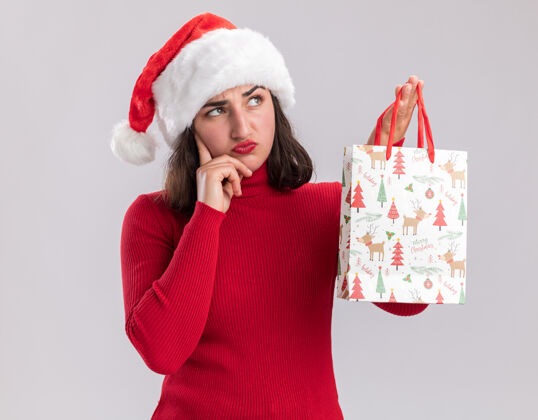圣诞老人穿着红色毛衣 戴着圣诞帽的小女孩手里拿着彩色纸袋和圣诞礼物 带着怀疑的表情站在白色背景下思考毛衣礼物帽子