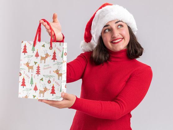 女孩快乐的年轻女孩穿着红色毛衣 戴着圣诞帽 手里拿着彩色纸袋和圣诞礼物 脸上挂着微笑 站在白色的背景下 望着一旁帽子微笑年轻人