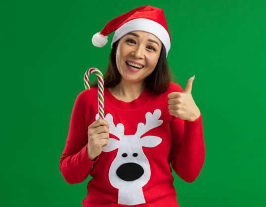 拇指年轻女子戴着圣诞老人帽 穿着红色毛衣 拿着糖果手杖 看着相机 开心地竖起大拇指站在绿色的背景上圣诞老人毛衣拿着