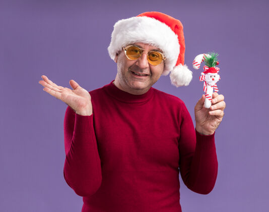 眼镜中年男子戴着圣诞帽戴着黄色眼镜拿着圣诞糖果手杖微笑着用手臂站在紫色的背景上呈现糖果手杖中间
