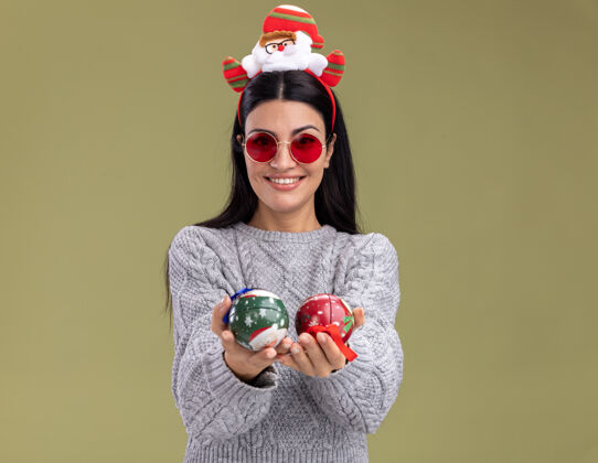 女孩微笑着的年轻白人女孩戴着圣诞老人的头带 戴着眼镜 把圣诞饰品伸向相机 看着相机孤立在橄榄绿的背景上 还有复印空间圣诞节头带眼镜