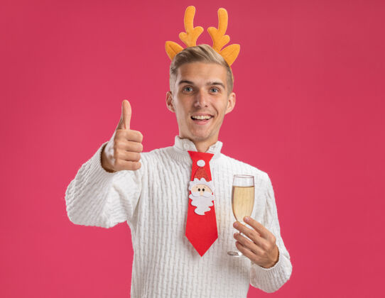 驯鹿快乐的年轻帅哥戴着驯鹿鹿角头带 打着圣诞老人的领带 手里拿着一杯香槟 大拇指向上竖起 孤立地站在粉色的墙上 留着复制空间领带穿着展示