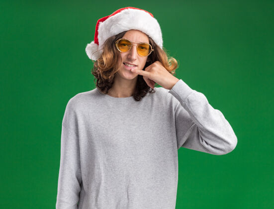 眼镜戴着圣诞老人帽戴着黄色眼镜的快乐的年轻人站在绿色的背景上看着摄像机做着叫我的手势圣诞老人年轻人做
