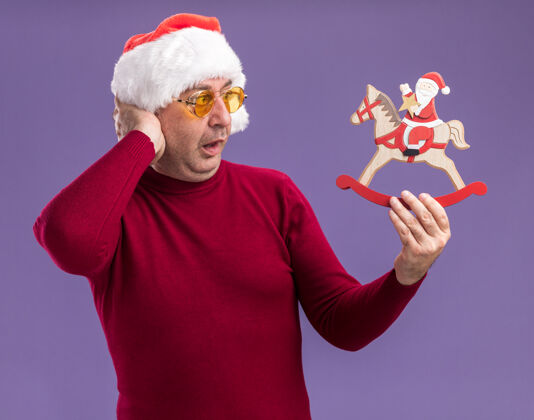 年龄中年男子戴着圣诞帽 戴着黄色眼镜 手里拿着圣诞玩具 站在紫色的背景下惊讶地看着它眼镜帽子中间