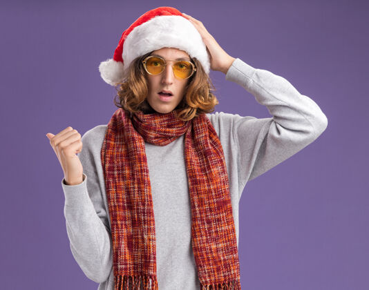 圣诞老人一个戴着圣诞老人帽 戴着黄色眼镜 脖子上围着暖和的围巾的年轻人惊讶地指着相机站在紫色背景上脖子围着帽子