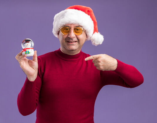 拿着快乐的中年男人戴着圣诞帽戴着黄色眼镜拿着圣诞雪球用食指指着它站在紫色的背景上圣诞老人年龄指着