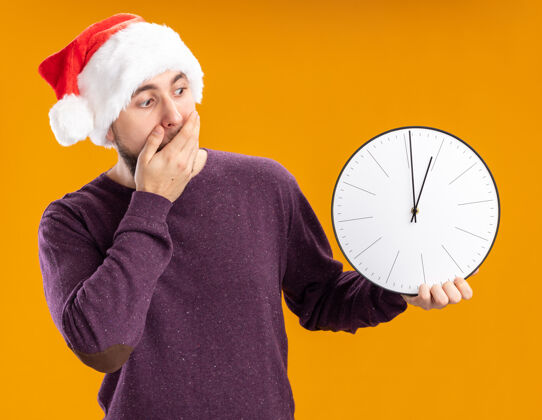 毛衣一个穿着紫色毛衣 戴着圣诞帽的年轻人拿着挂钟看着它 惊讶地用手捂住嘴巴站在橙色的背景上拿着圣诞老人钟