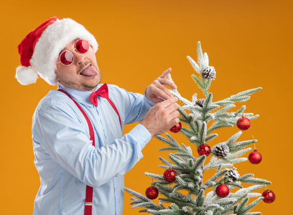 蝴蝶结快乐的年轻人戴着吊带领结 戴着圣诞帽 戴着红眼镜 站在圣诞树旁 把玩具挂在树上 高兴又惊讶地伸出舌头 遮住橙色的背景领带悬挂圣诞节