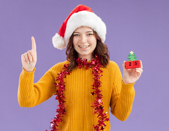 装饰品微笑着的年轻斯拉夫女孩 戴着圣诞帽 脖子上戴着花环 手拿圣诞树饰物 在紫色背景上孤立地向上指 还有复制空间微笑圣诞指着