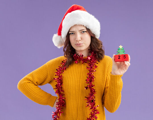 圣诞树失望的年轻斯拉夫女孩戴着圣诞帽 脖子上戴着花环 手拿圣诞树饰物 紫色背景上有复制空间周围女孩花环
