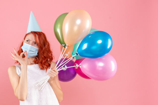 举行前视图中的年轻女子拿着五颜六色的气球在无菌面具上粉红色的墙壁气球病毒年轻女性