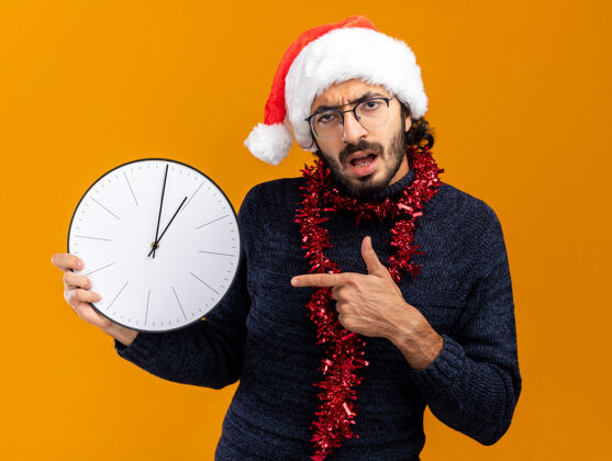 未出租年轻帅哥戴着圣诞帽 脖子上戴着花环 指着橙色背景的挂钟举行墙壁圣诞节