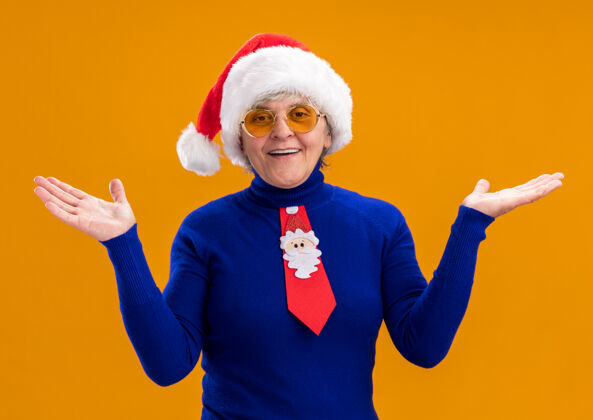 微笑戴着太阳眼镜 戴着圣诞帽 系着圣诞领带的微笑的老妇人 手拉着手 在橙色的背景上与复制空间隔离开来眼镜手老人