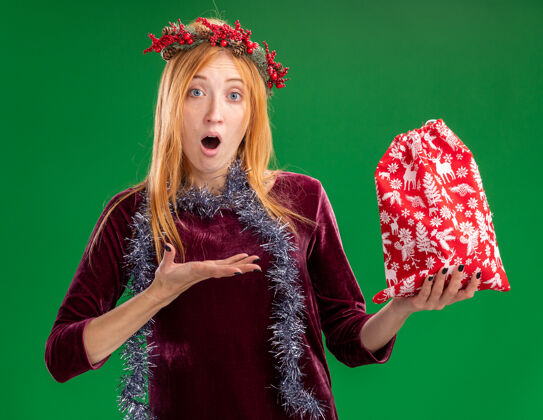 点年轻漂亮的女孩穿着红色的裙子 脖子上戴着花环和花环 手指着绿色背景上的圣诞包女孩惊喜手