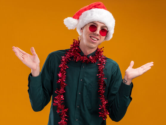 镀金快乐的年轻金发男子戴着圣诞帽 戴着眼镜 脖子上戴着金箔花环 两手空空地看着孤立在橙色背景上的摄像机空快乐脖子